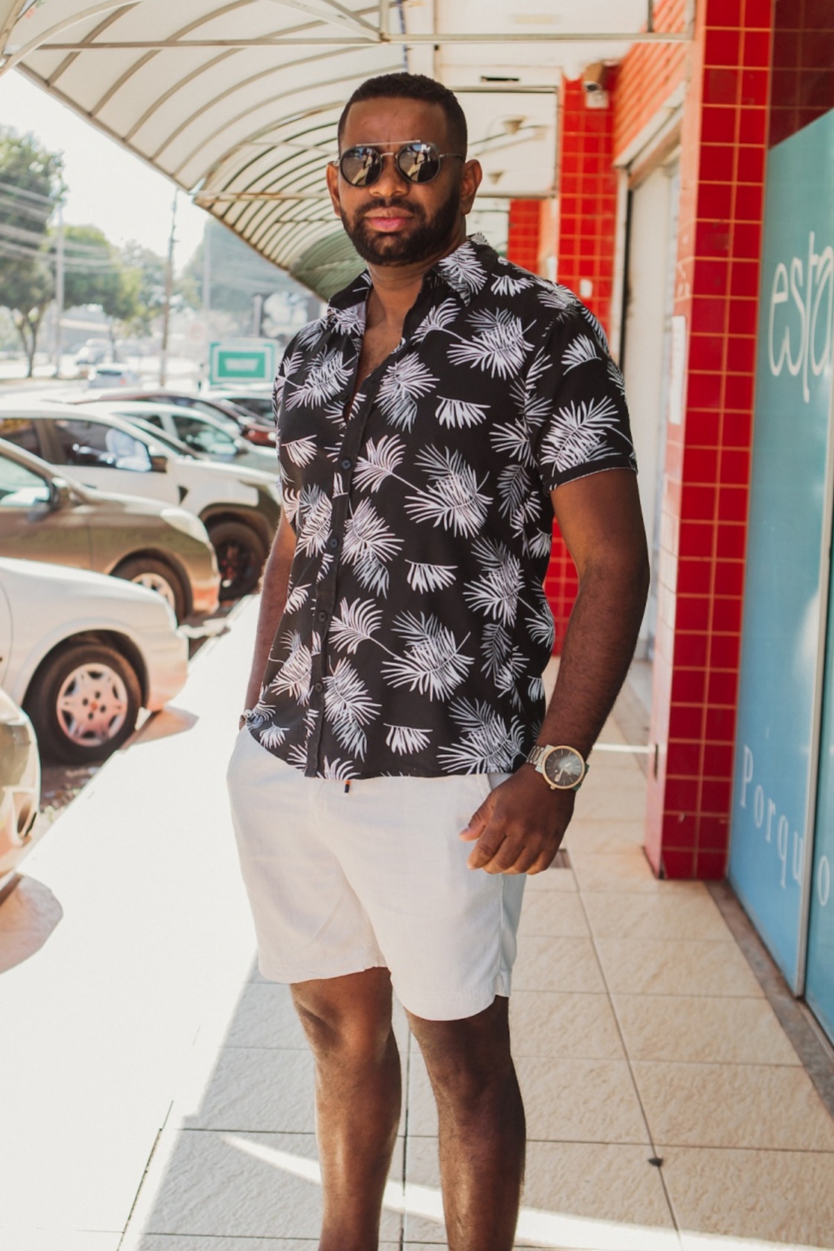 Homem posa para a foto. Com uma camisa de botão florida em preto e branco ele olha pra a câmera com seus óculos escuros e bermuda branca.