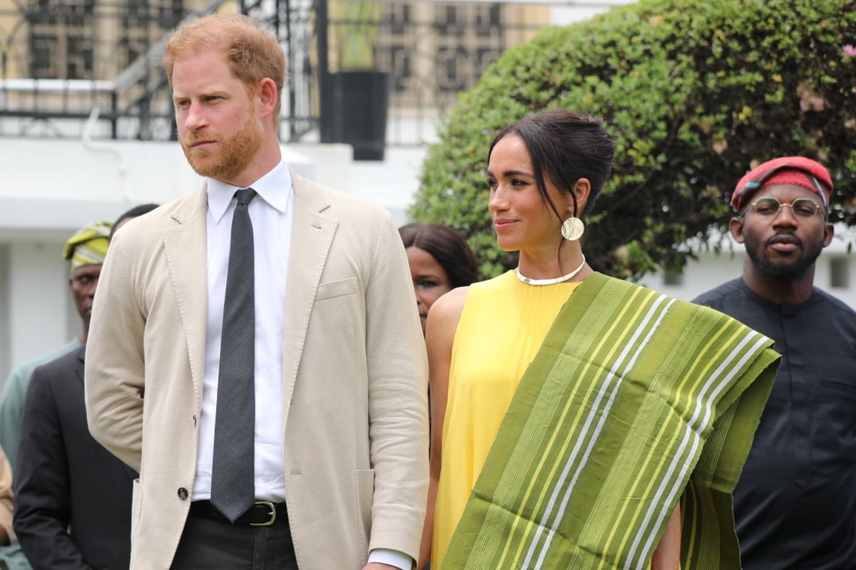 Foto colorida de homem com blazer bege ao lado de mulher com vestido amarelo e echarpe verde. Harry e Meghan Markle - Metrópoles