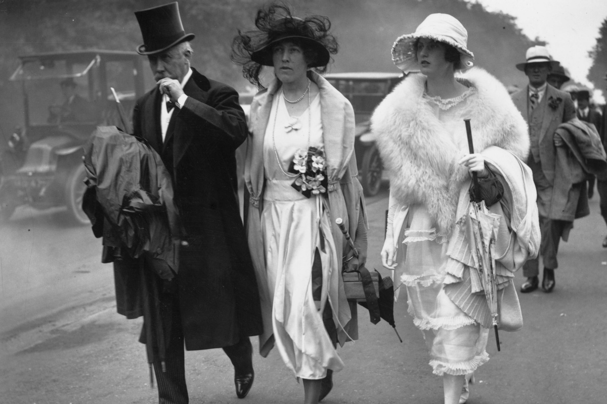 Da esquerda para a direita. Um homem e duas mulheres caminham para evento. A imagem, fotografada em preto e branco, mostra o trio andando com suas roupas clássicas e seus acessórios.