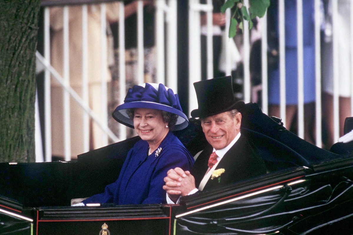 Rainha Elizabeth e Príncipe Philip posam para a foto. A rainha veste um visual todo em roxo, junto a um chapéu que acompanha as peças. O Príncipe veste um smoking preto com uma cartola sob a cabeça. Ambos estão sentados em um carro