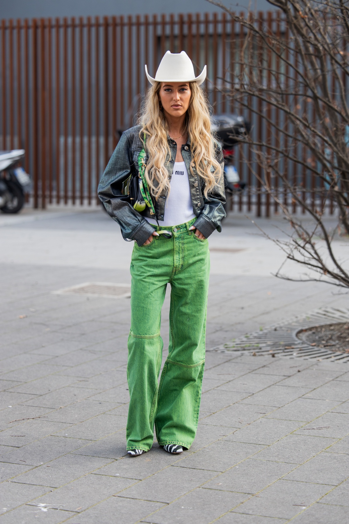 Mulher posa para a foto. Com uma calça verde, ela combina seu visual com uma jaqueta cinza e uma camiseta regata branca. Como acessório, ela usa um chapéu branco sob a cabeça