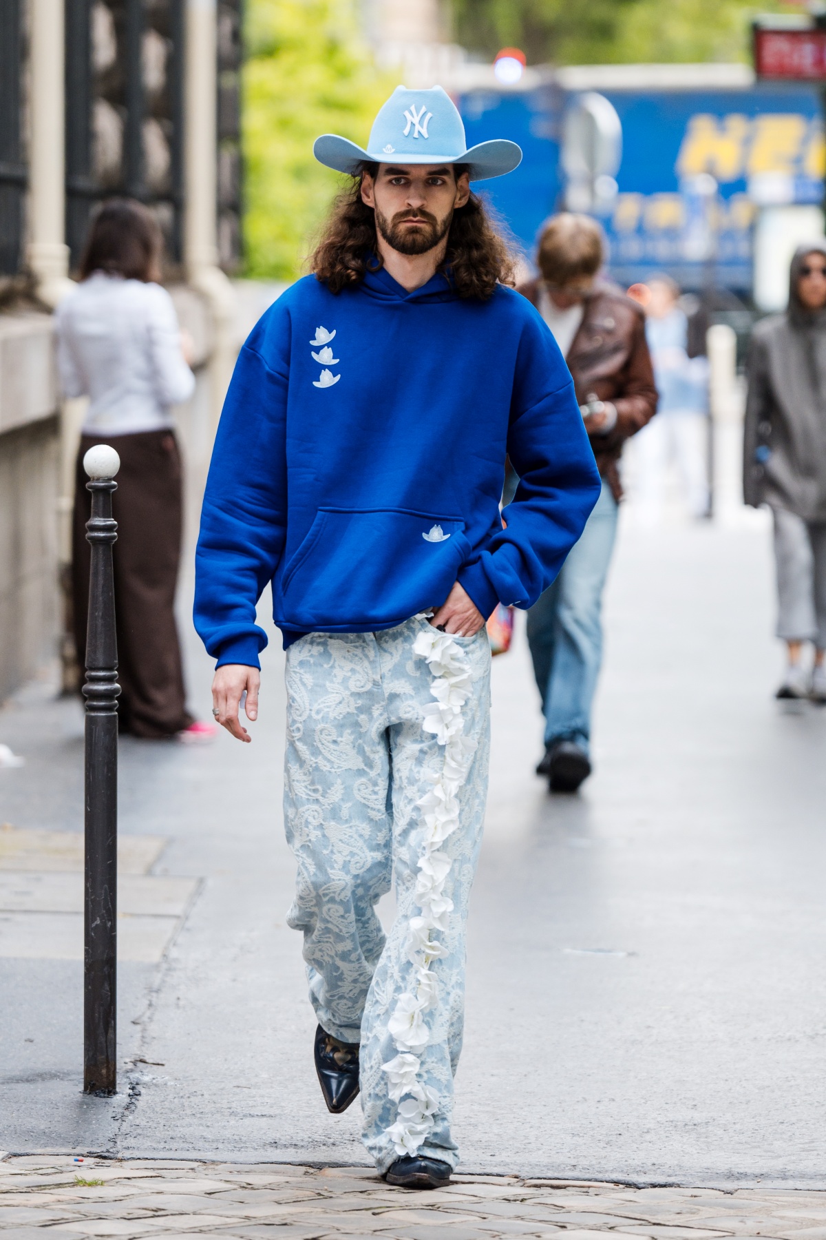 Homem posa para a foto. Com o visual majoriatiamente em azul e branco, ele veste um tênis de corrida, calça jeans e um casaco moletom. Como acessório, o homem usa um chapéu azul sob a cabeça.