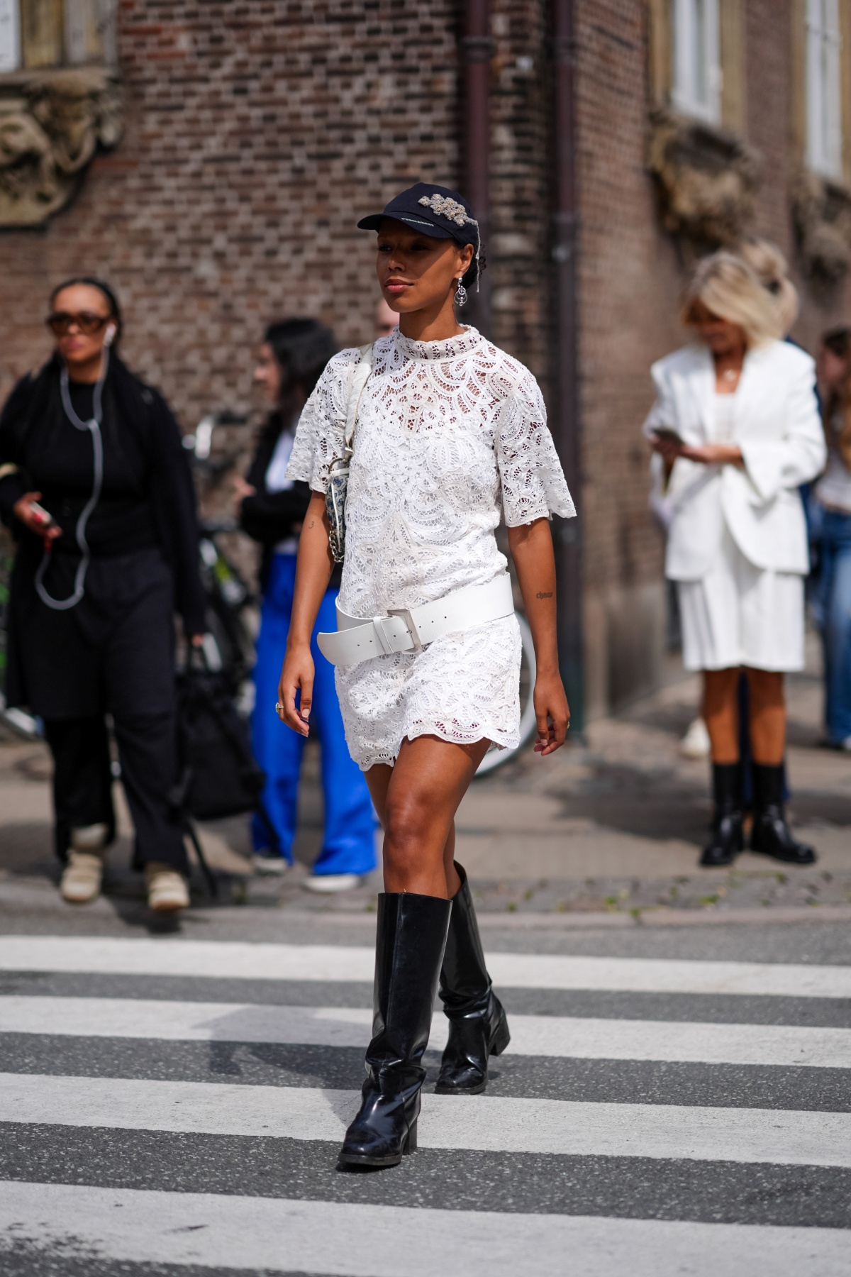 Mulher posa para a foto. Atravessando a rua, ela veste um conjunto branco de bermuda e camiseta, junto à uma boata preta, combinando com um boné, também preto.