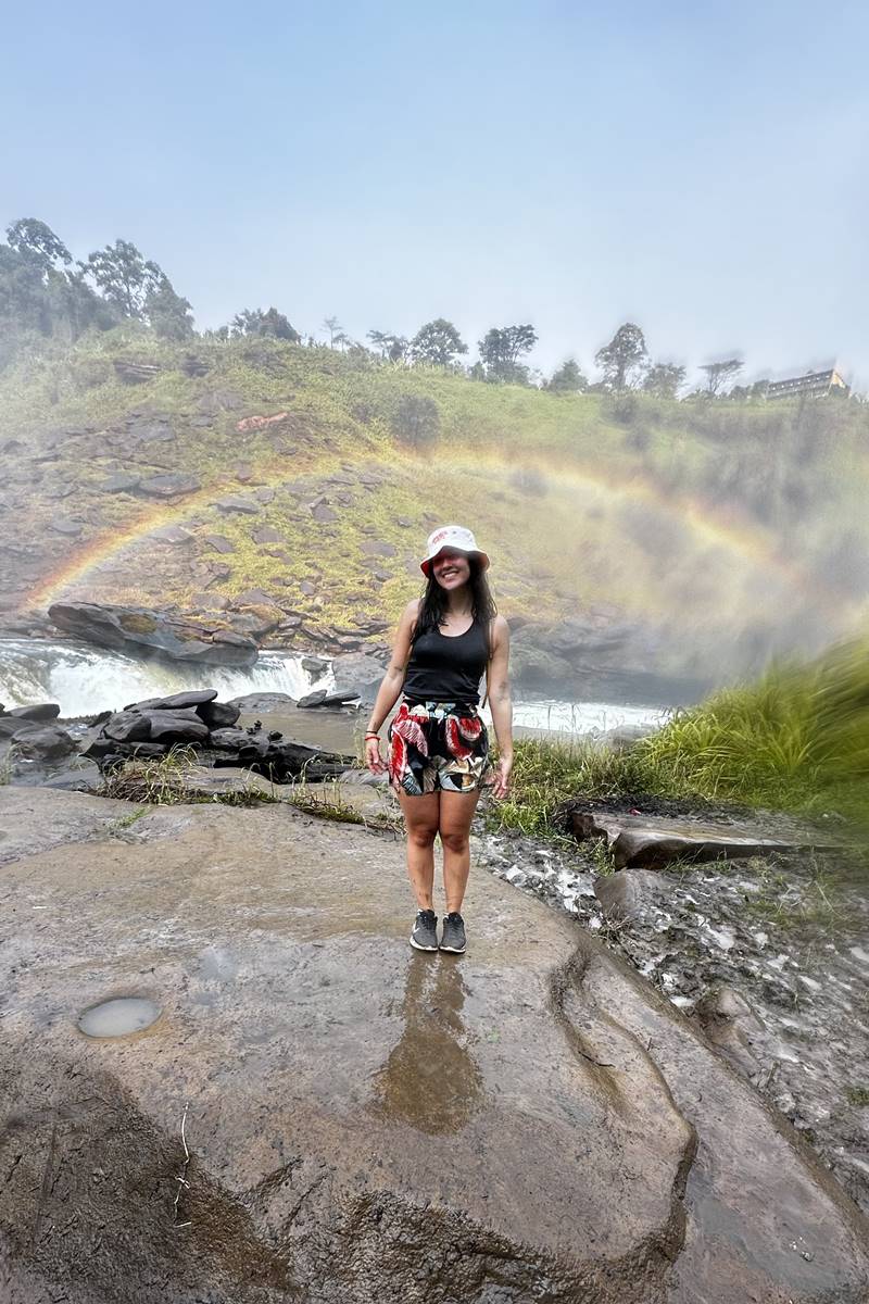 Foto colorida de mulher em cachoeira com arco-íris - Metrópoles