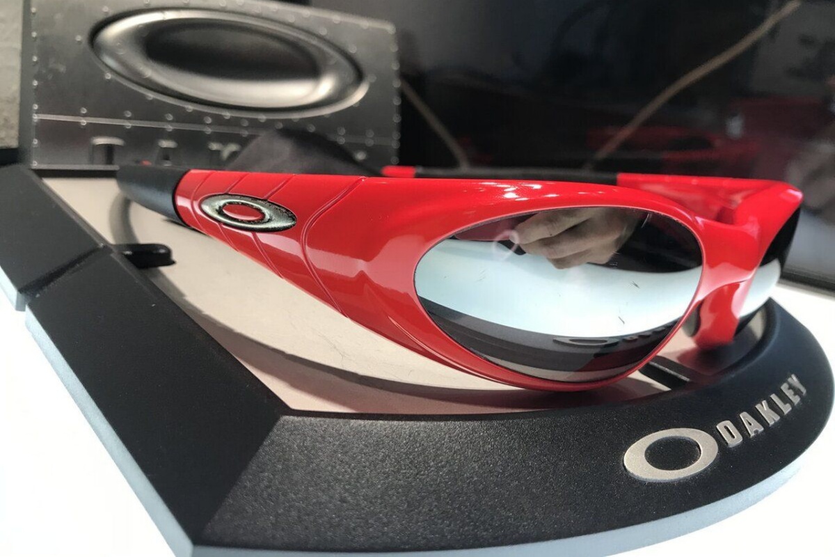 A imagem mostra um óculos vermelho e preto sob uma base da marca Oakley