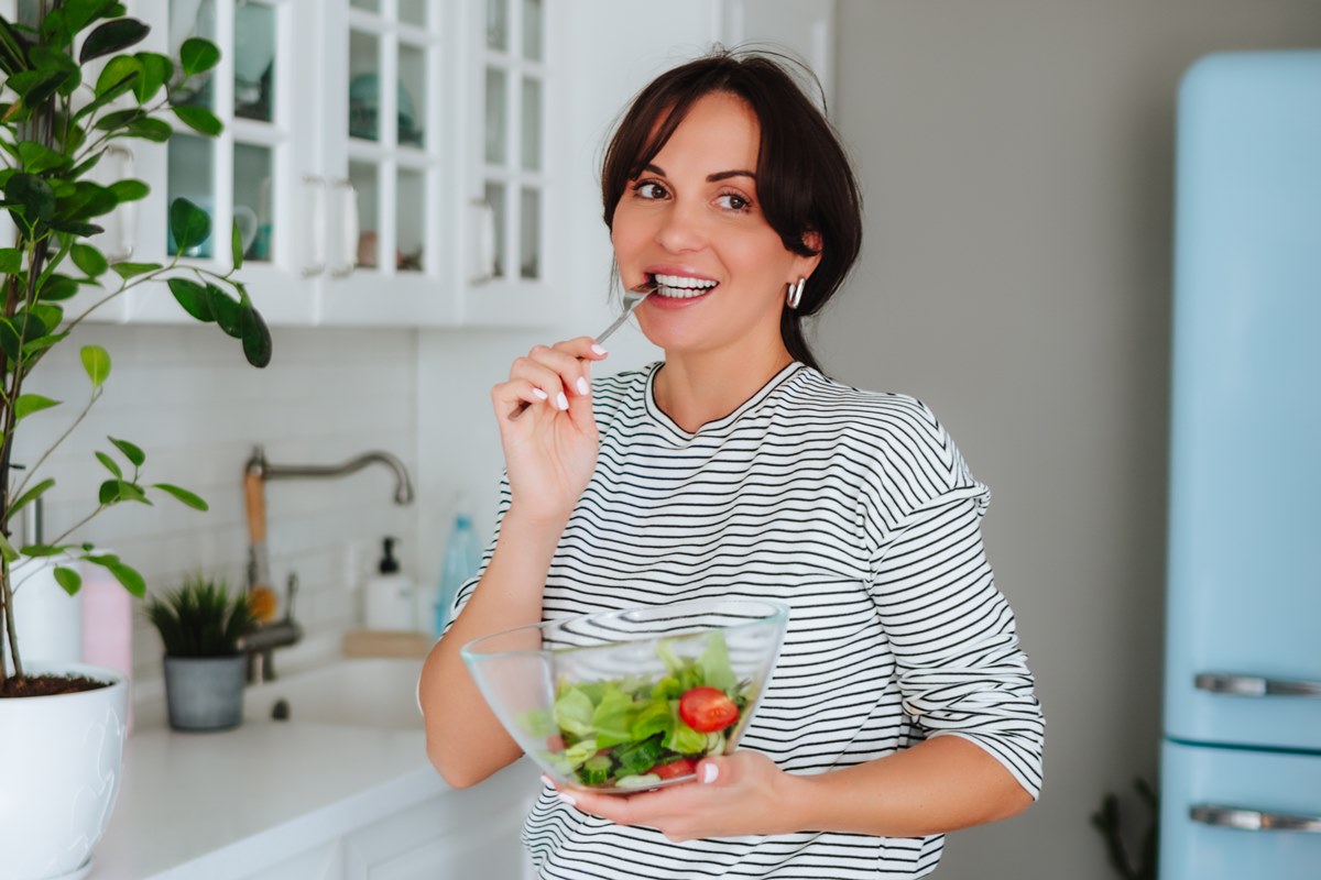 Foto colorida de mulher em cozinha comendo uma salada - Metrópoles