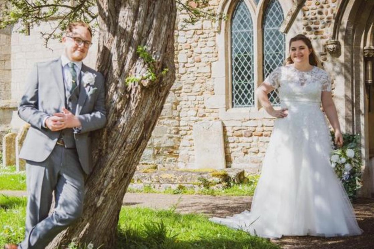 Foto do casal Nathan e Katia no dia do casamento. O homem está de terno encostado em uma arvore, e a noiva mais atrás sorrindo -Metrópoles