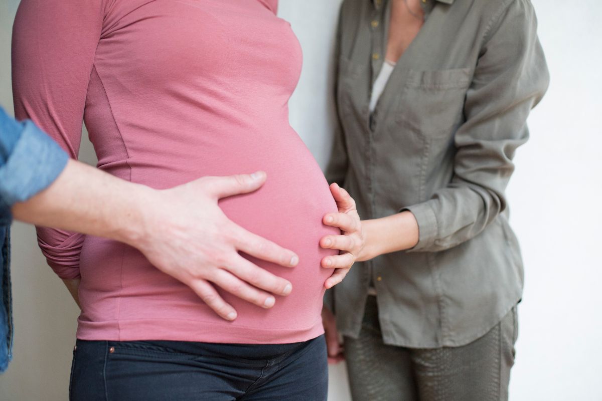 Foto colorida de uma mulher grávida e duas pessoas com a mão na barriga - Metrópoles