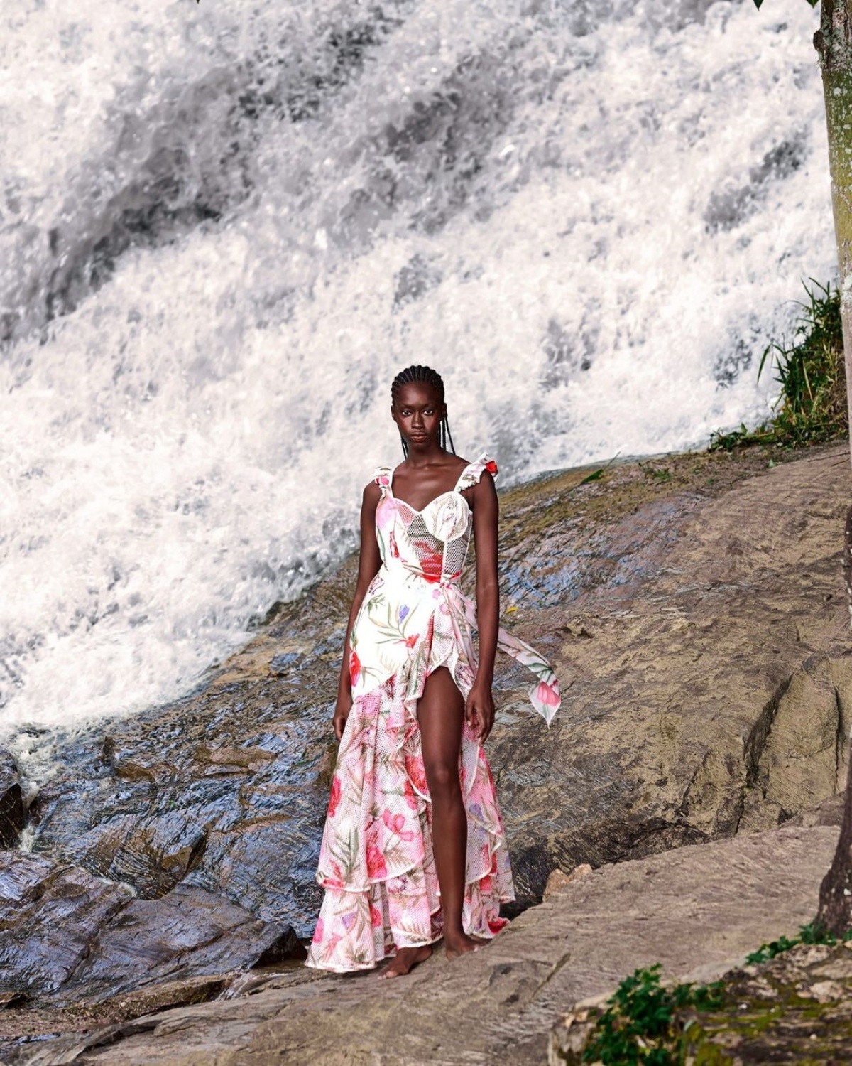 Mulher posa para a foto em um cenário de natureza. Em frente à uma cachoeira, ela olha fixamente para a foto enquanto usa um vestido branco com detalhes vermelhos