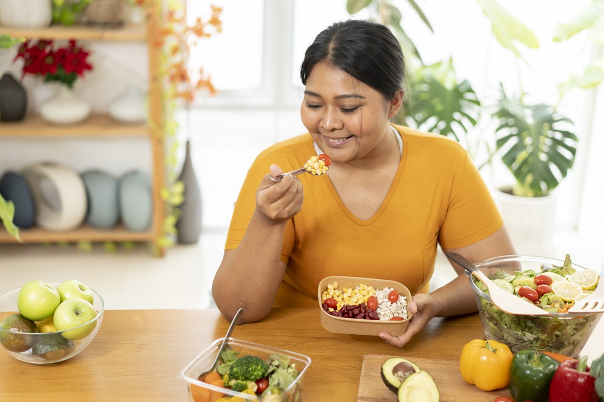 Foto colorida de mulher sentada comendo salada. Ela está em uma mesa com legumes e verduras - Metrópoles