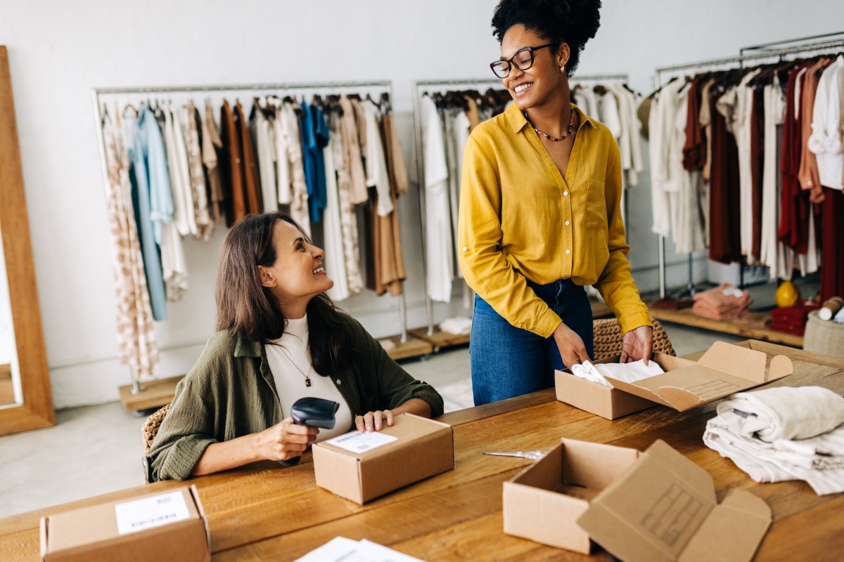 A imagem mostra duas mulheres de negócios sorrindo umas para as outras enquanto atendem pedidos feitos em sua loja de roupas online.