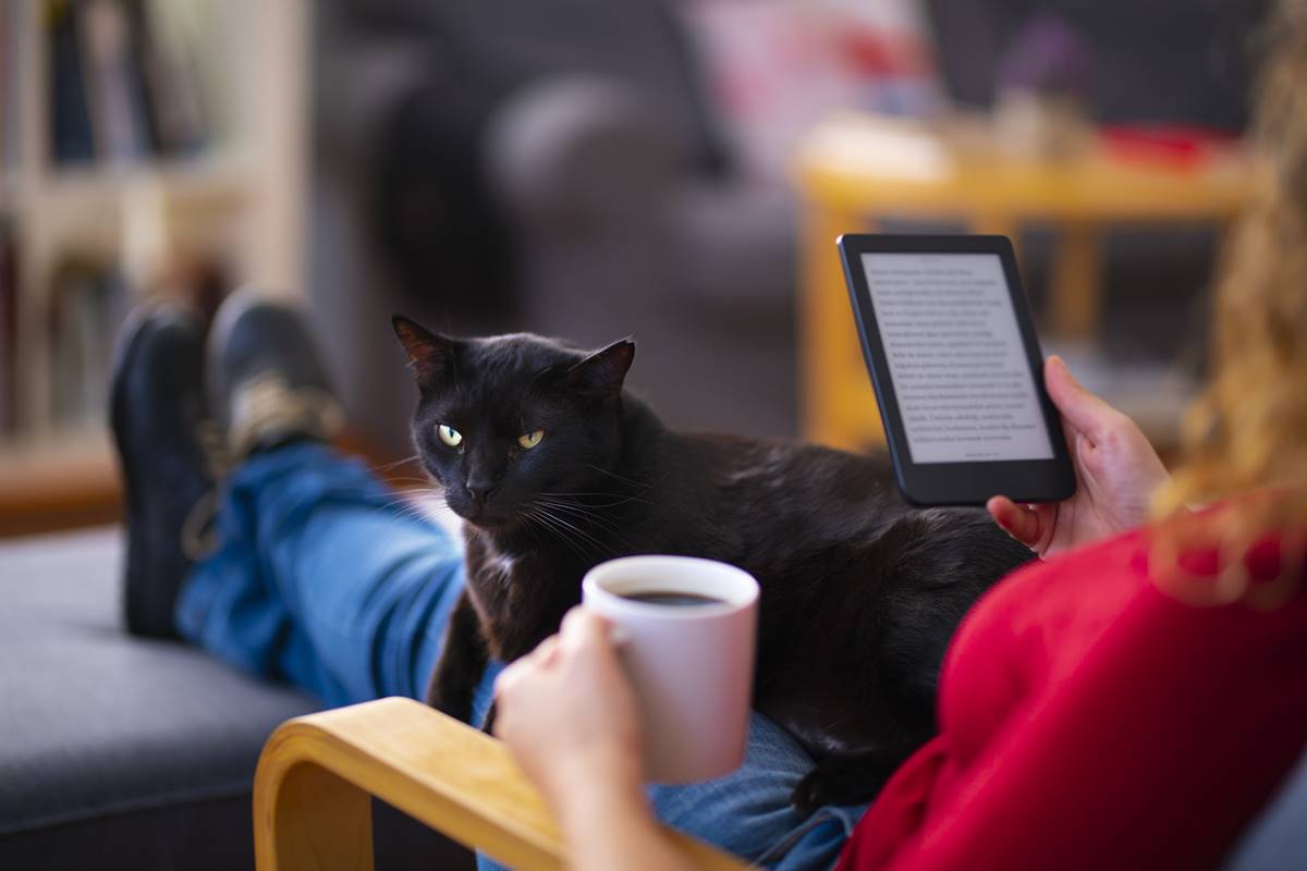 Mulher sentada lendo livro digital, enquanto segura caneca de café. Gato preto aparece deitado no colo da mulher - Metrópoles