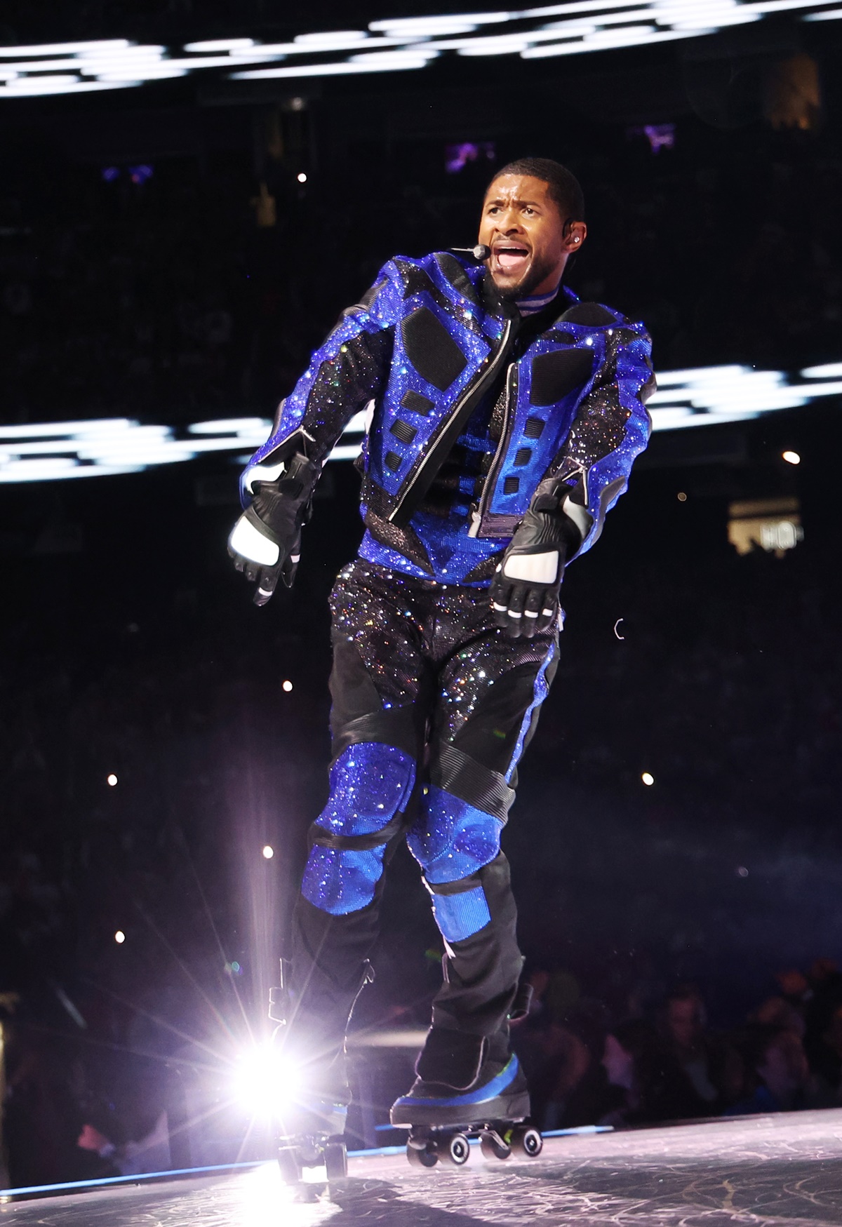 A imagem mostra o cantor Usher vestindo uma conjunto azul e preto cobreto de brilho enquanto patina no palco