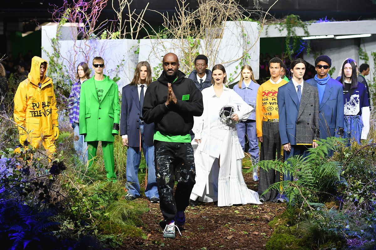 A imagem mostra o designer Virgil Abloh finalizando seu desfile. Atrás do estilista estão seus modelos vestindo roupas sociais nas cores verde, amarelo, azul e branco.