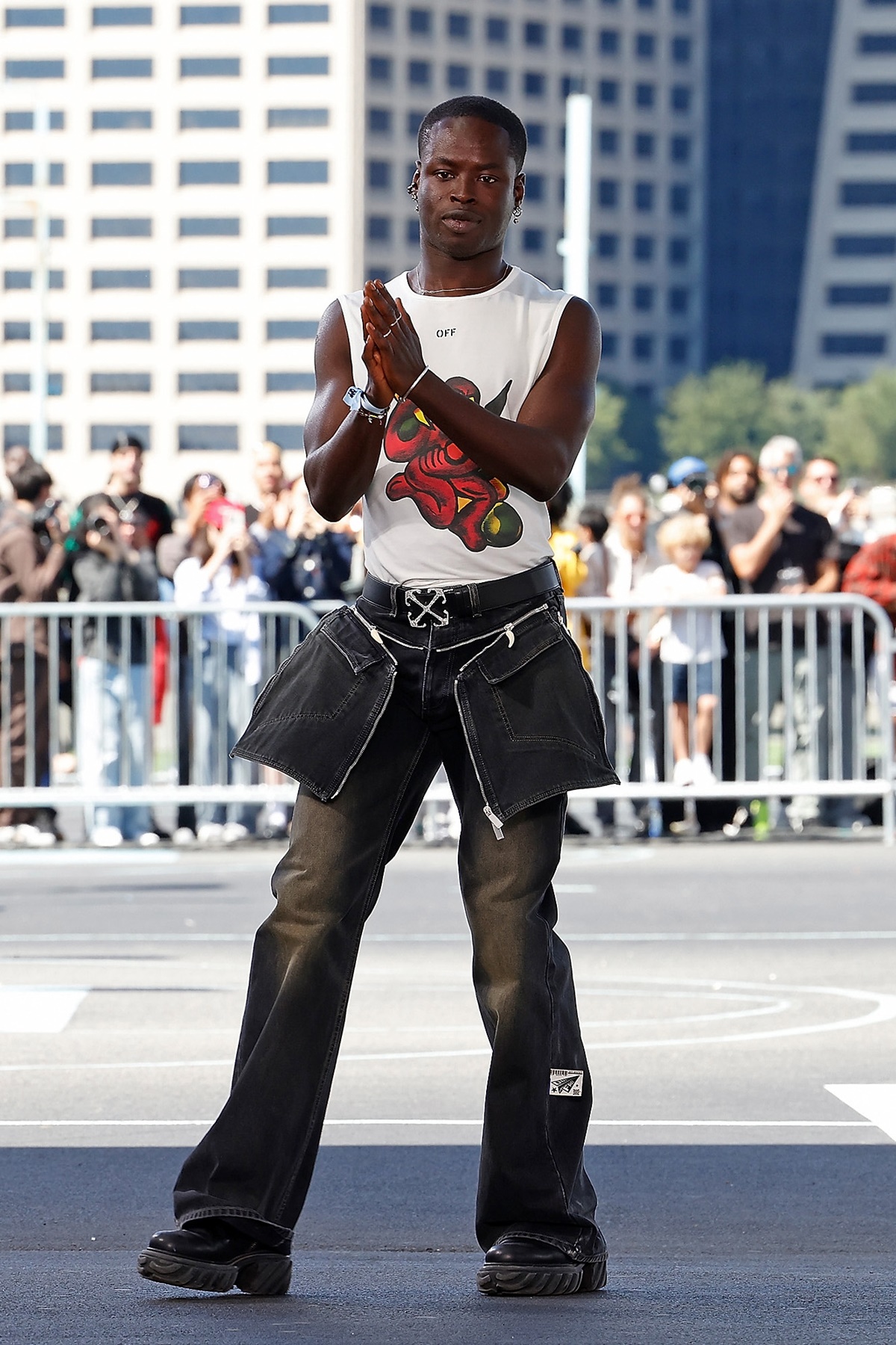 A imagem mostra o designer Ibrahim Kamara após o desfile. Calçando um coturno preto e calças jeans, o jovem designer veste uma camiseta com um estampa vermelha e mãos postas