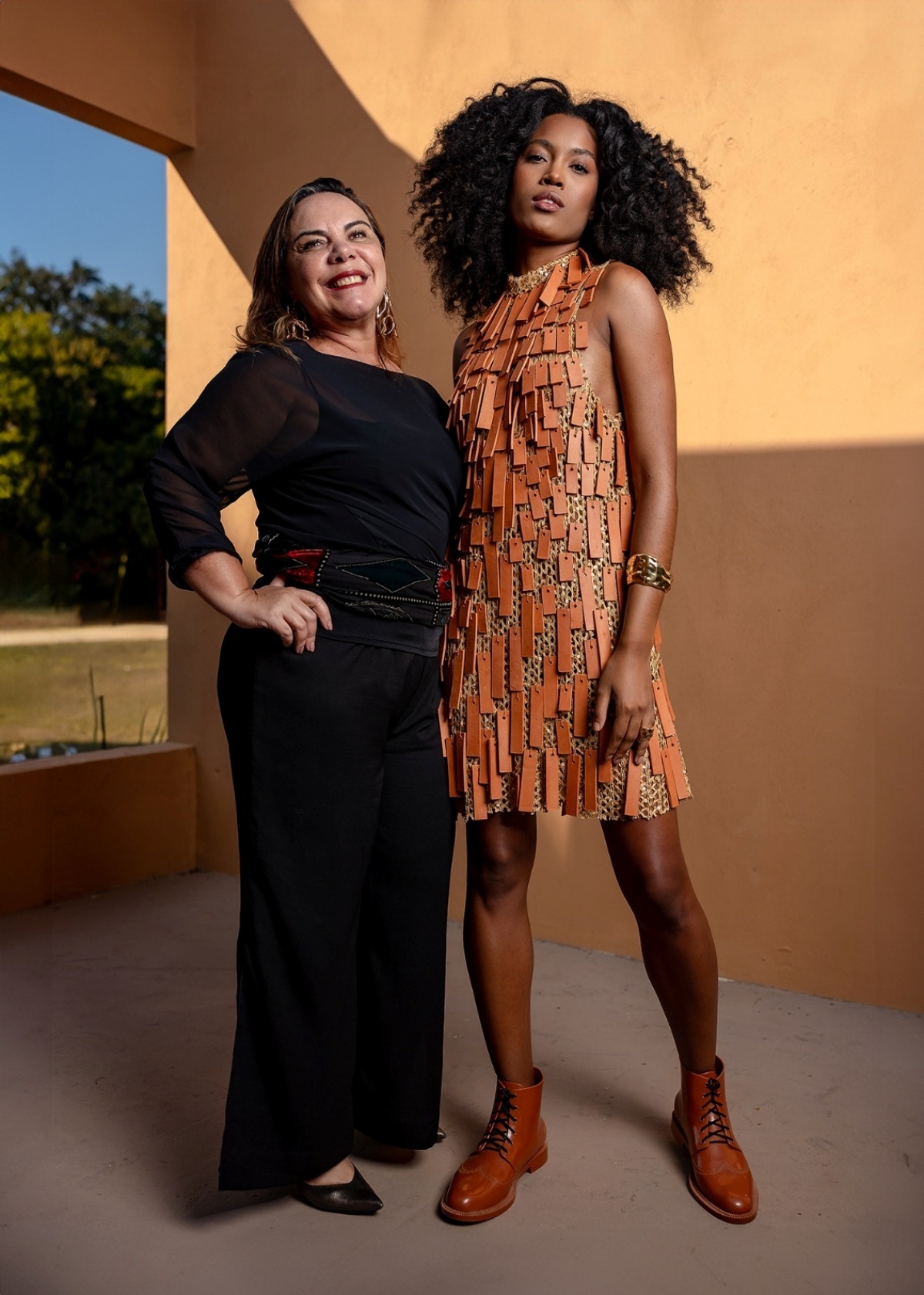 Mulheres posam para a foto. A modelo à direita da imagem posa olhando para a câmera com seu vestido em retalhos marrons euanto a outra à esquerda olha para o horizonte com seu visual completamente preto