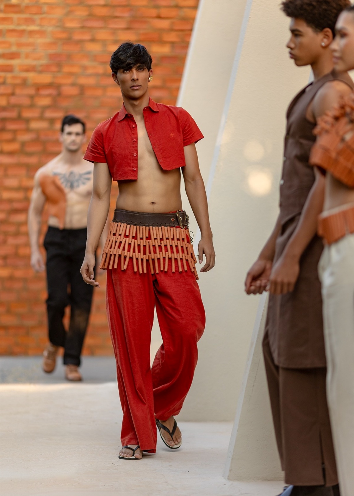 A imagem mostra um modelo desfilando. Vestindo um conjunto de camisa e calça vermelha, ele caminha com acessórios na cintura