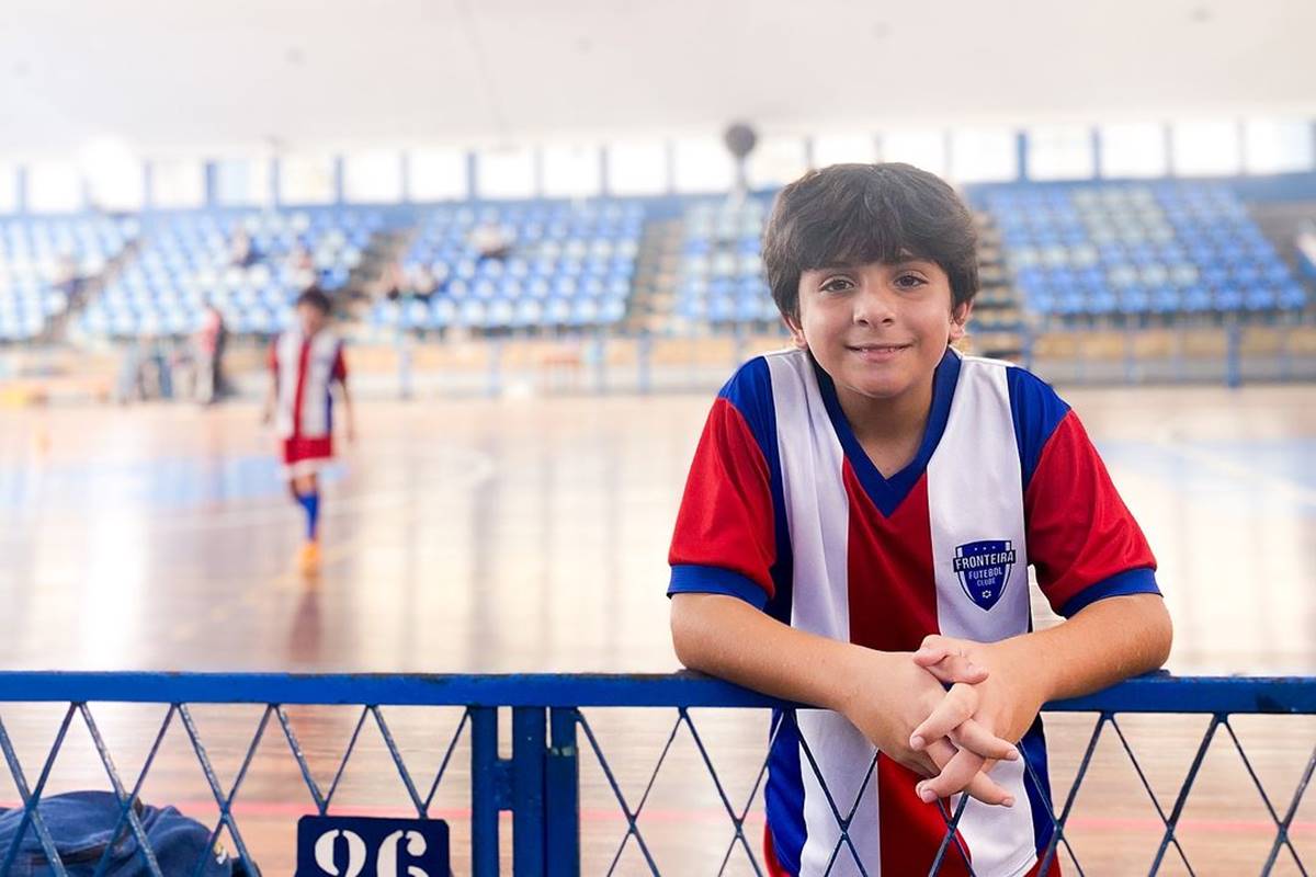 foto colorida de menino de uniforme de futebol posando apoiado em barreira de metal - metrópoles