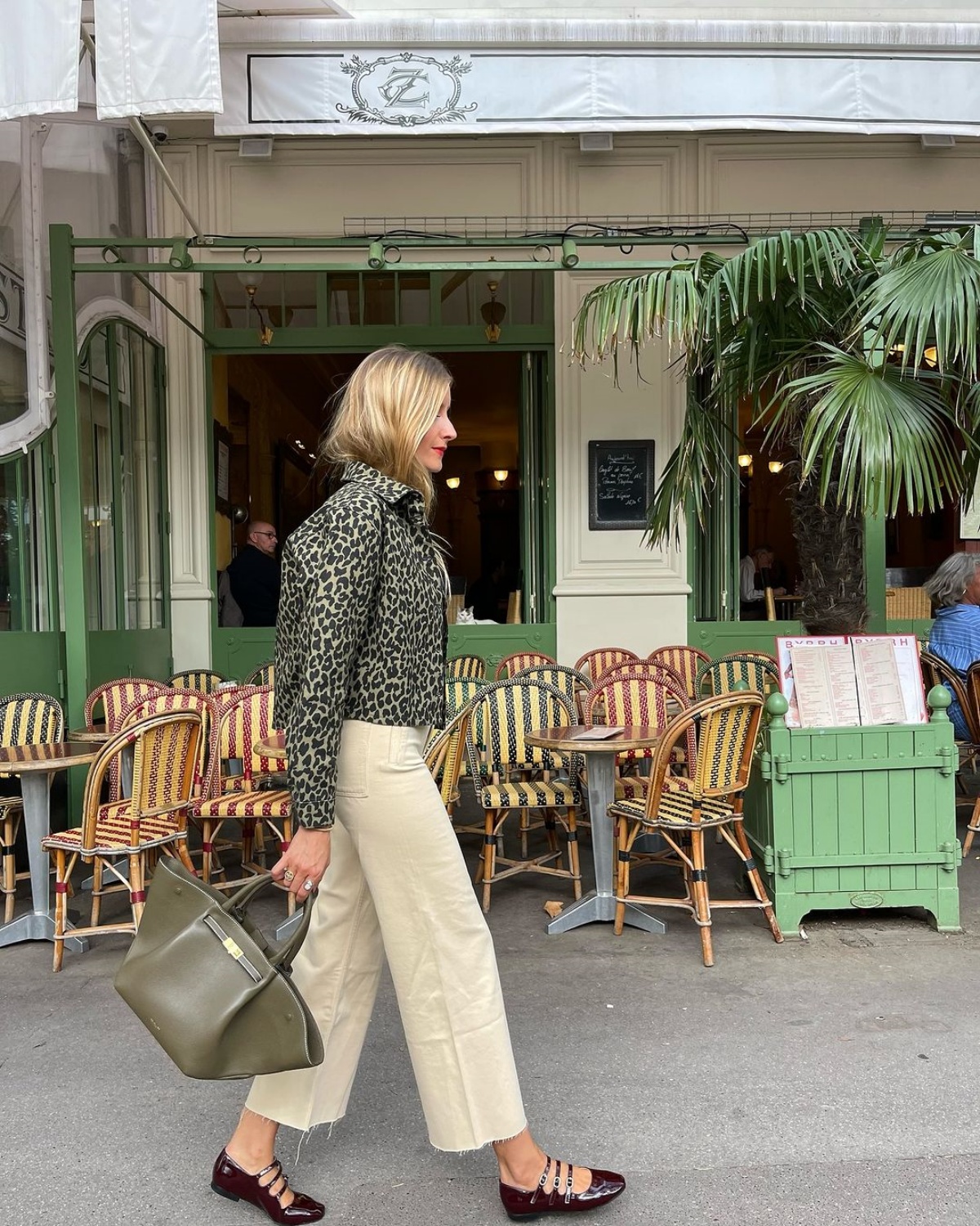 A imagem mostra uma mulher caminhando em frente uma lanchonete. Vestindo um casaco com estampa animal, ela usa uma calça bege com uma bolsa verde e sapatilha vermelha