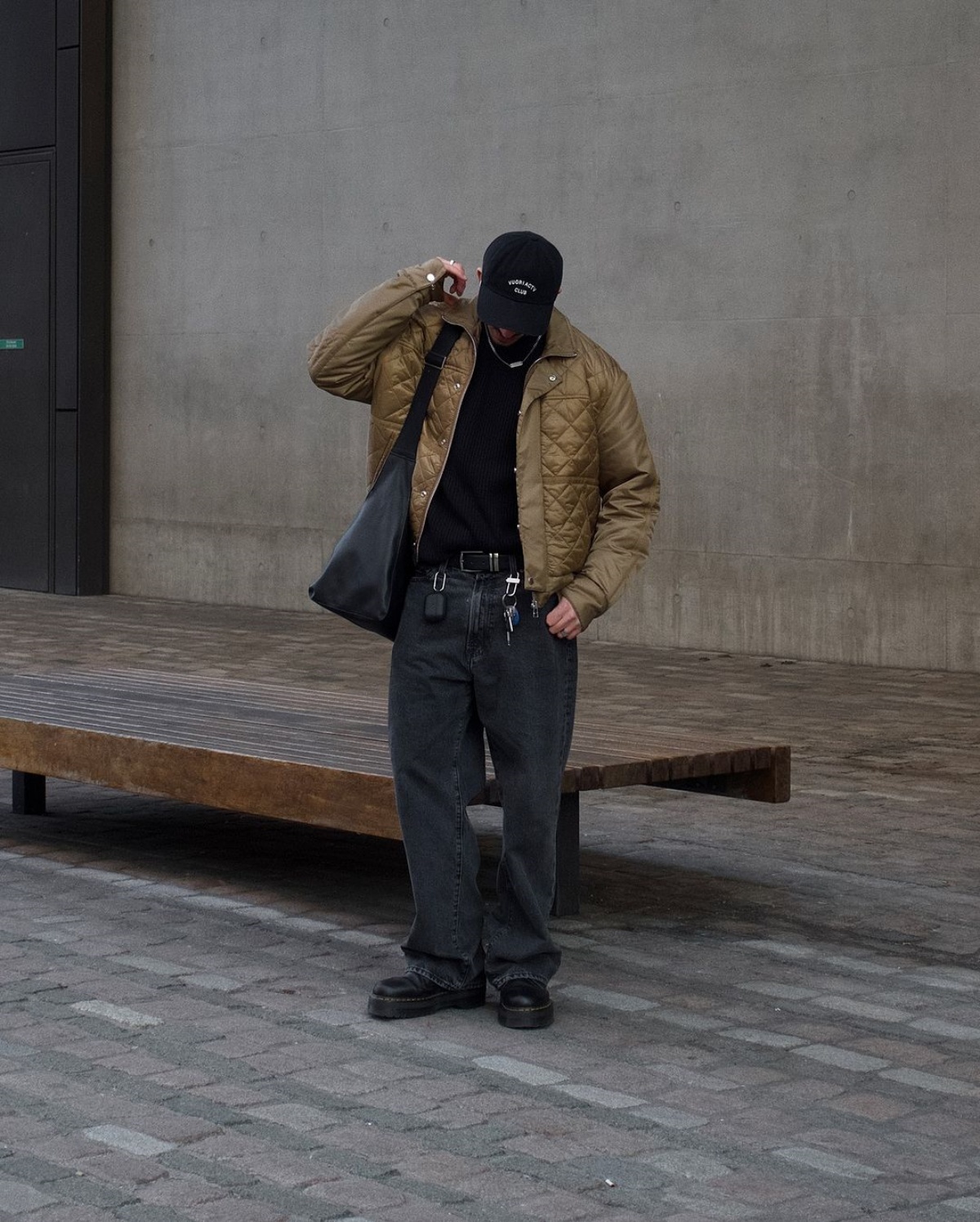 A imagem mostra um homem com um boné preto, jaqueta verde e camiseta preta carregando uma bolsa. Nos pés ele calça uma bota preta combinando com sua calça preta desbotada