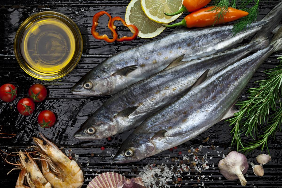 Foto colorida de três peixes cavalinha em uma superfície de madeira. Ao lado, tem pimentão cortado, azeite e folhas verdes - Metrópoles