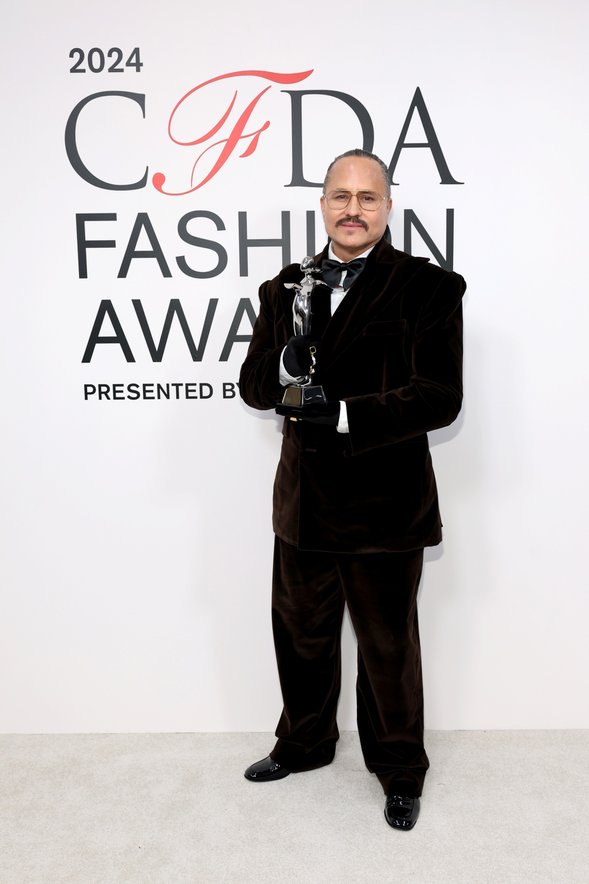 A imagem mostra o estilista Willy Chavarria empunhando seu troféu em frente ao backdrop do CFDA Fashion Awards