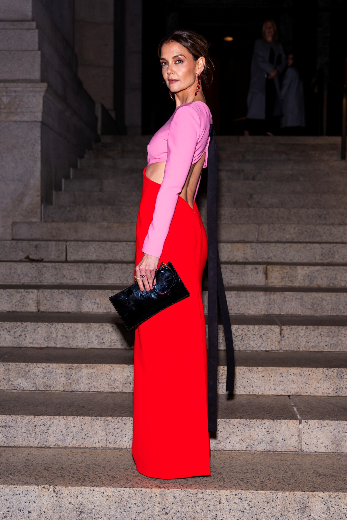 A imagem mostra a atriz Katie Holmes fotografada em frente uma escadaria. Usando um vestido nas cores rosa e vermelho, Holmes carrega um bolsa preta na mão esquerda que combina com um laço preto nas costas