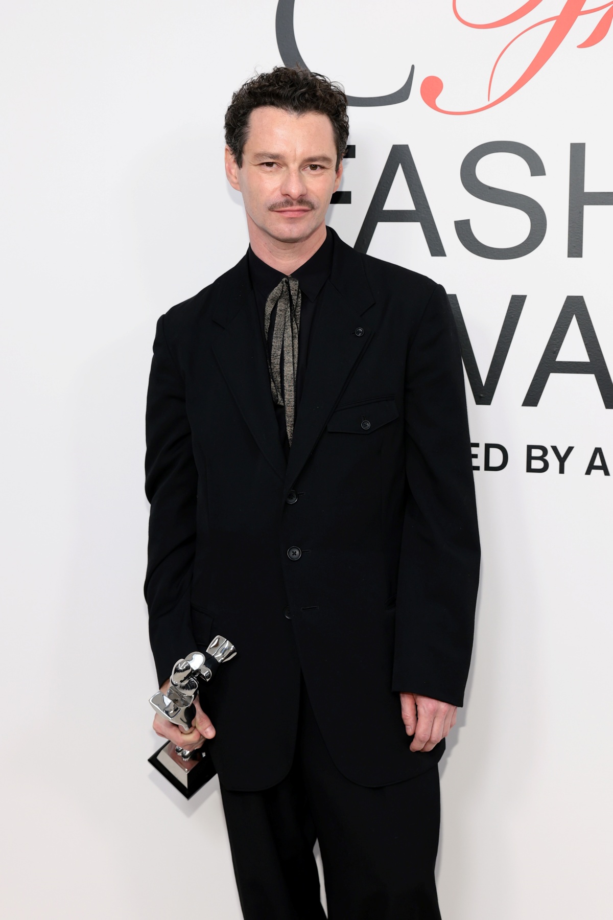 A imagem mostra o designer Henry Zankov em frente ao backdrop do CFDA Fashion Awards empunhando seu troféu