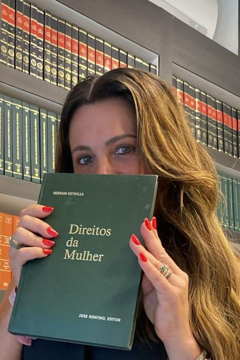 Foto colorida de mulher em biblioteca com a capa de um livro na frente do rosto - Metrópoles