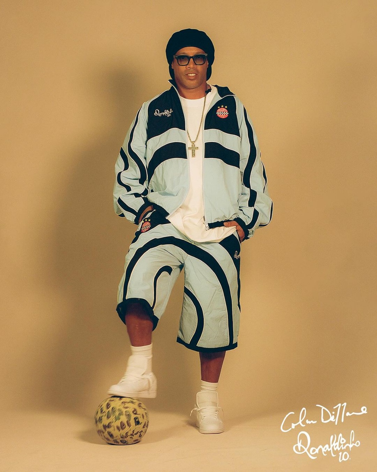 A imagem morta o jogador Ronaldinho Gaúcho posando para a foto. Ronaldinho veste um conjunto de casaco e bermuda azul com detalhes pretos.