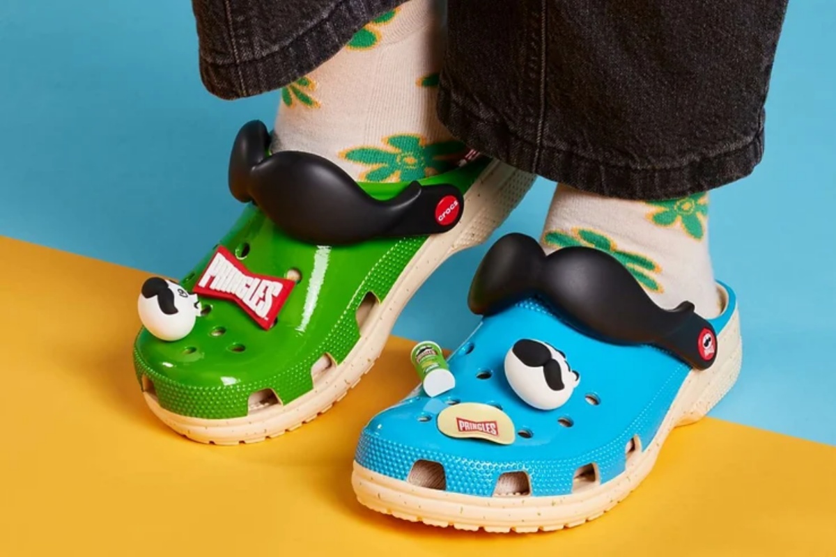 A imagem mostra um par de Crocs. O pé esquerdo na cor azul e o direito na cor verde. Ambos trazem um bigode como detalhe na frente