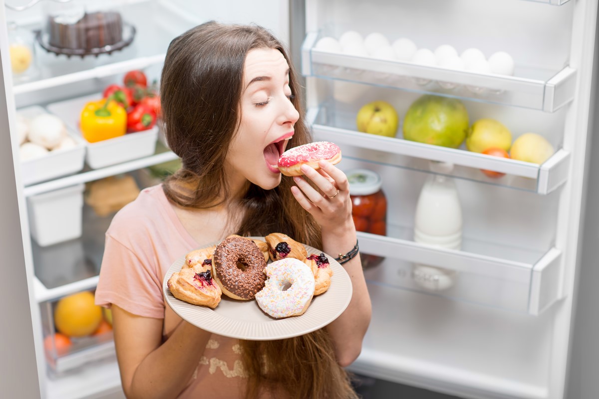 Foto colorida de mulher sentada entre as portas de uma geladeira e com um prato com donuts. Ela está comendo um doce - Metrópoles