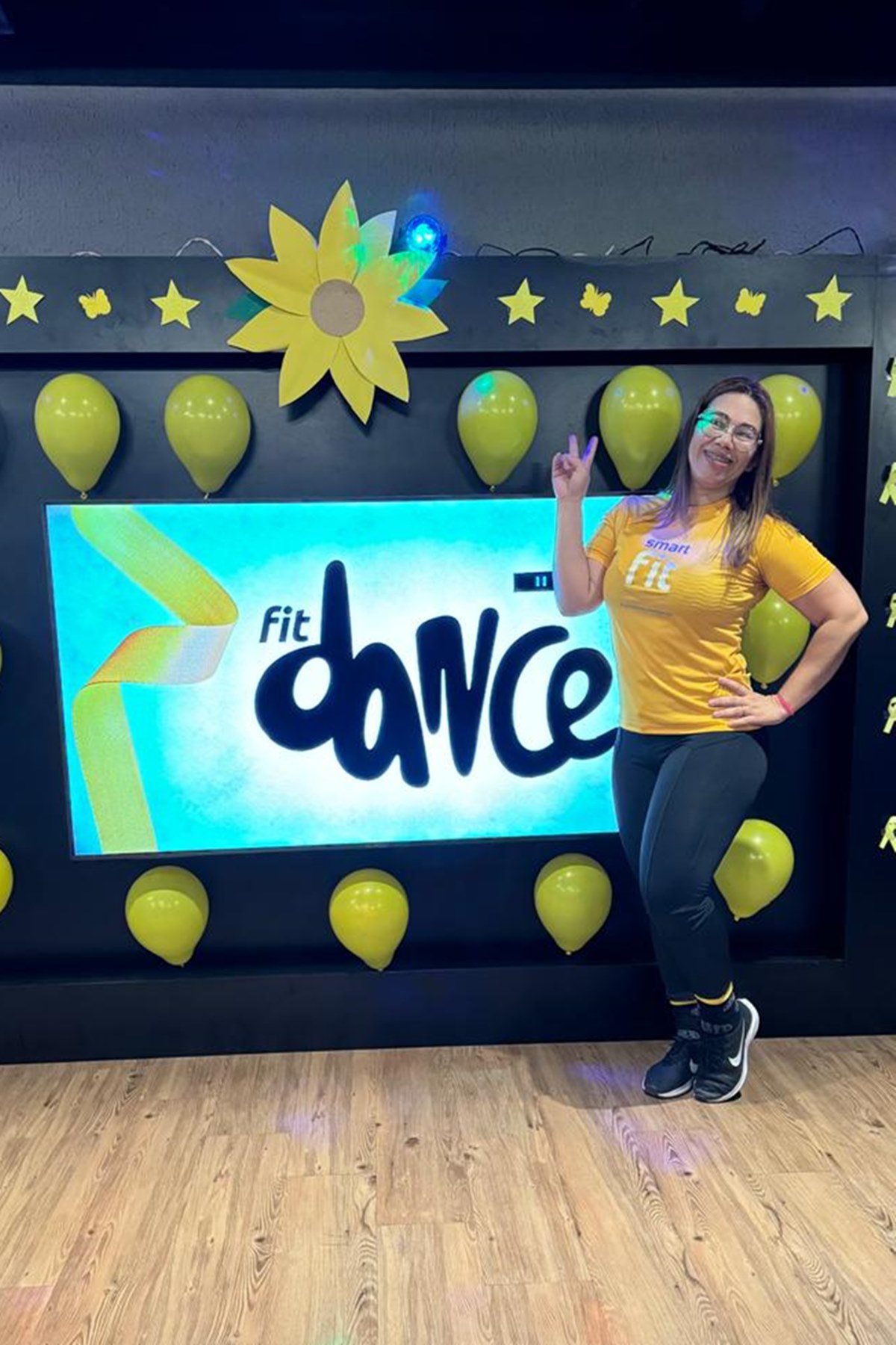 Imagem mostra Gizelle Ribeiro Valadares, uma mulher branca, de blusa amarela, posando em frente a um poster sobre fit dance - Metrópoles