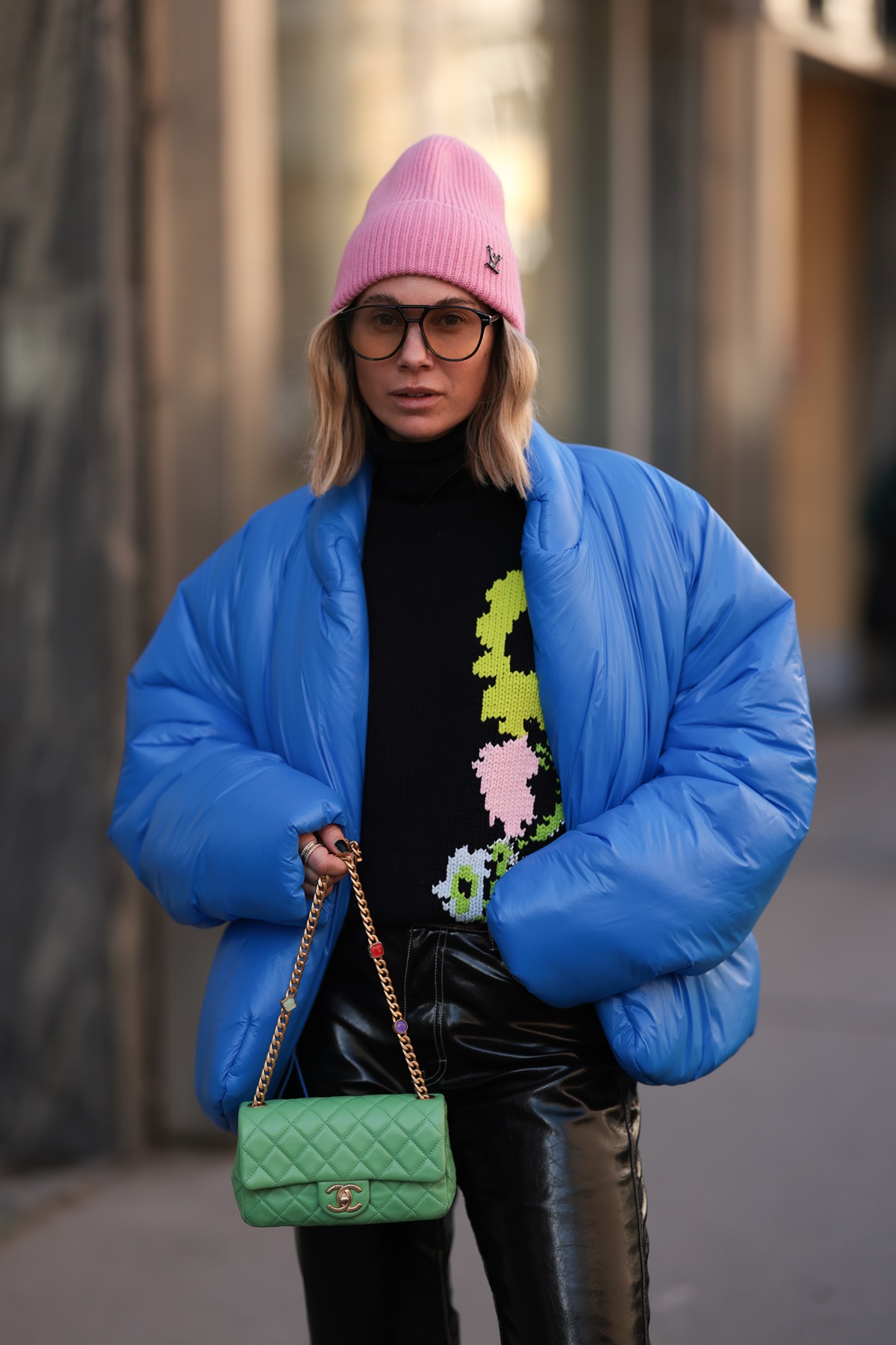 A imagem mostra uma mulher vestindo uma touca rosa, camiseta preta combinando com um casaco azul e uma bolsa verde