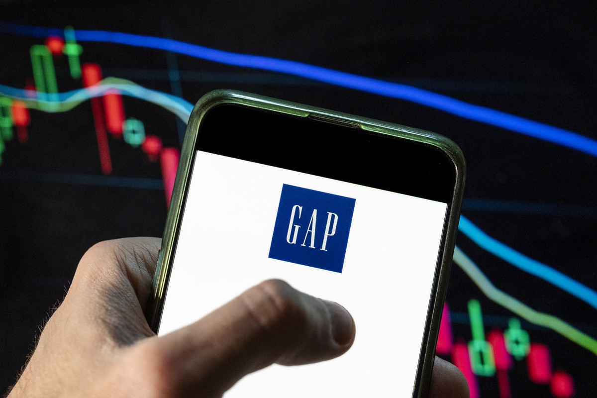 A imagem mostra uma mão segurando um celular com a logo da Gap em frente à uma tela com gráficos em queda