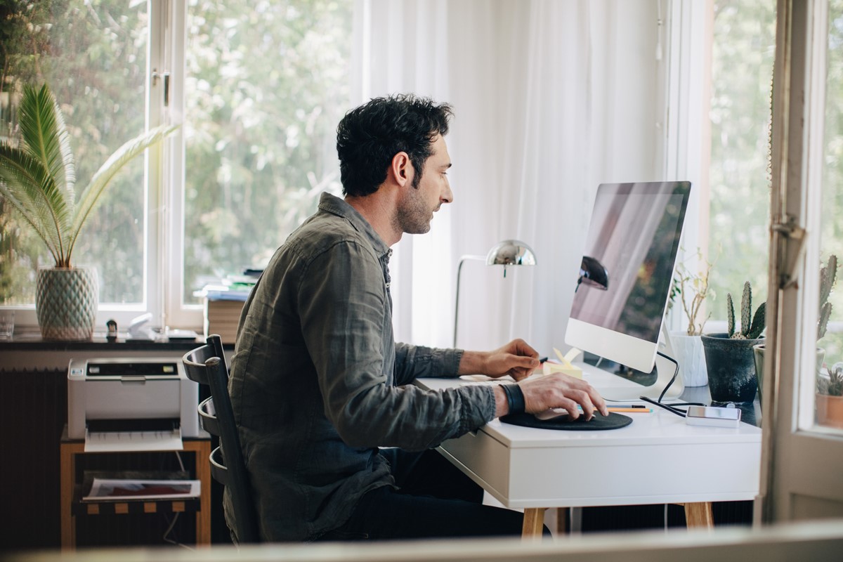 Vista lateral do jovem empresário usando o computador enquanto está sentado na mesa no escritório em casa. Metrópoles