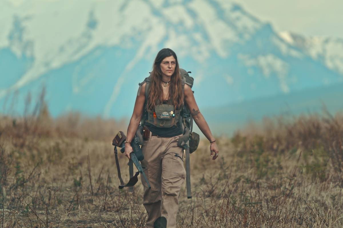 Imagem colorida mostra mulher forte, segurando bolsa e armas, andando em descampado - Metrópoles