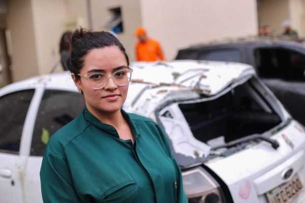 Foto colorida de mulher morena de costas para carro destruído por arvore caída