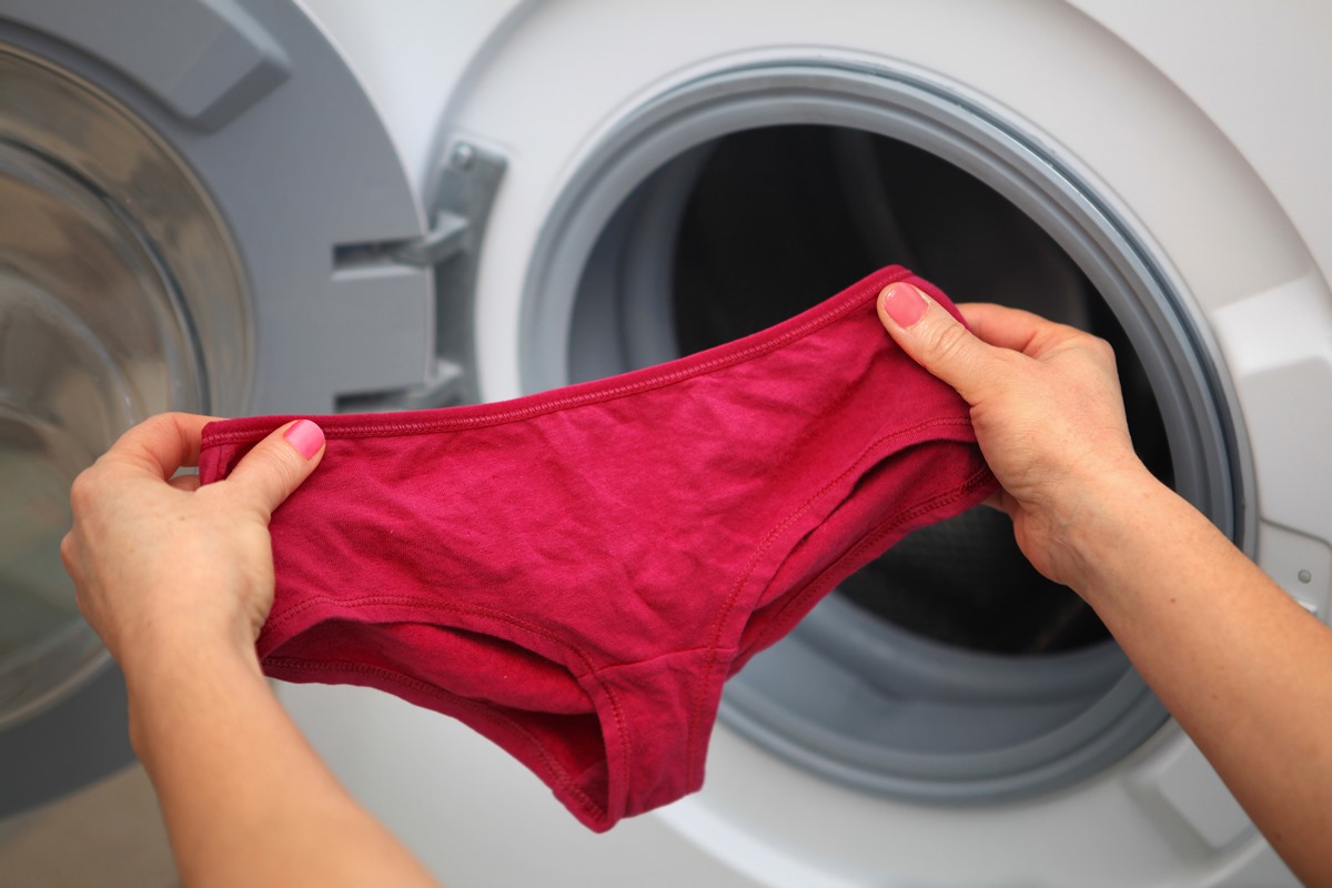 Foto colorida de mãos de uma mulher segurando uma calcinha perto de uma máquina de lavar - Metrópoles