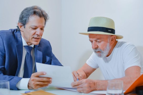 Imagem colorida mostra homem branco de terno e gravata azuis segurando folha de papel. Ele está sentado em uma mesa ao lado de homem idoso, com barba branca, camiseta branca e chapéu na cabeça - Metrópoles