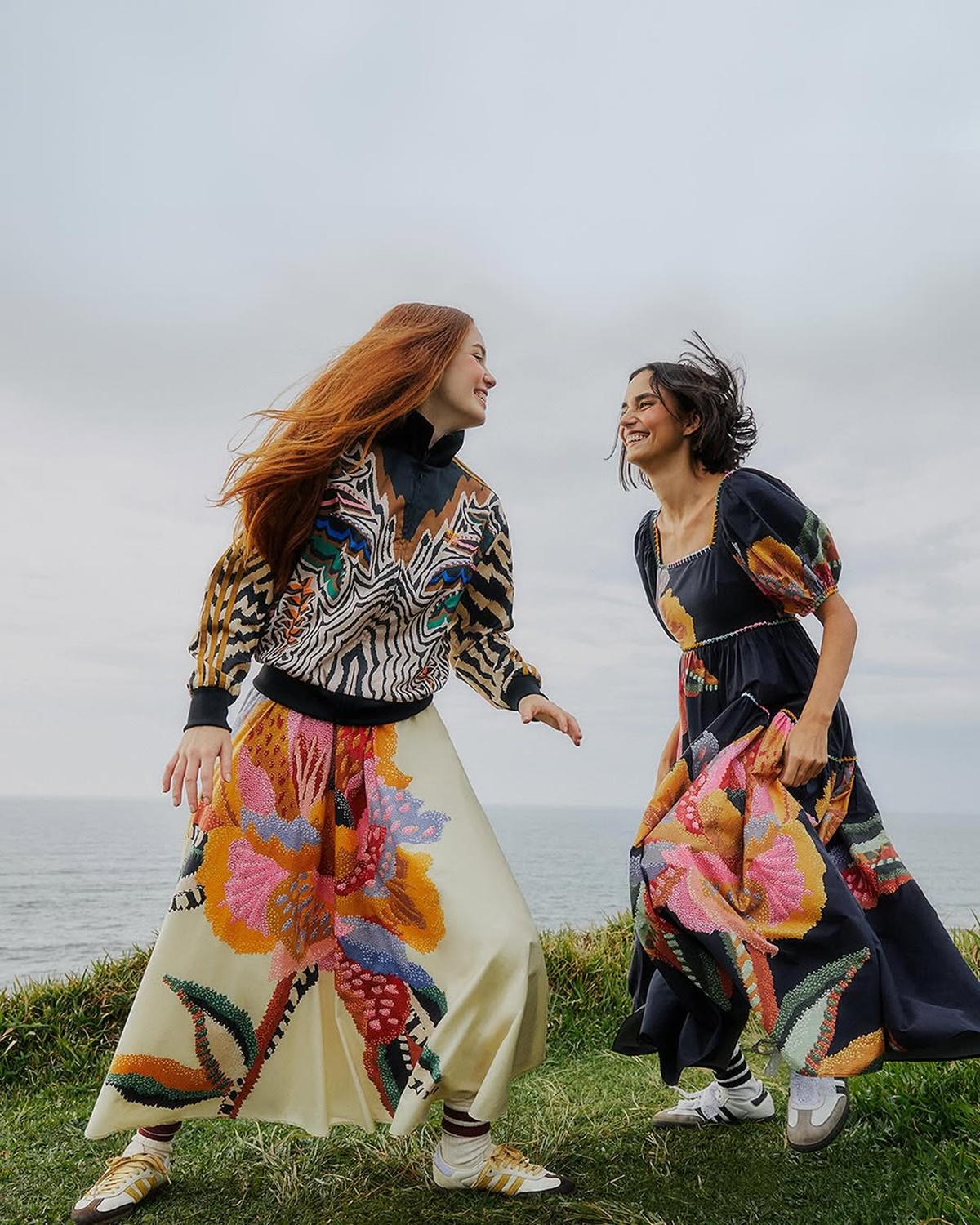 A imagem mostra duas modelos felizes. Vestindo roupas coloridas, ambas sorriem enqnato andam em um ambiente arborizado