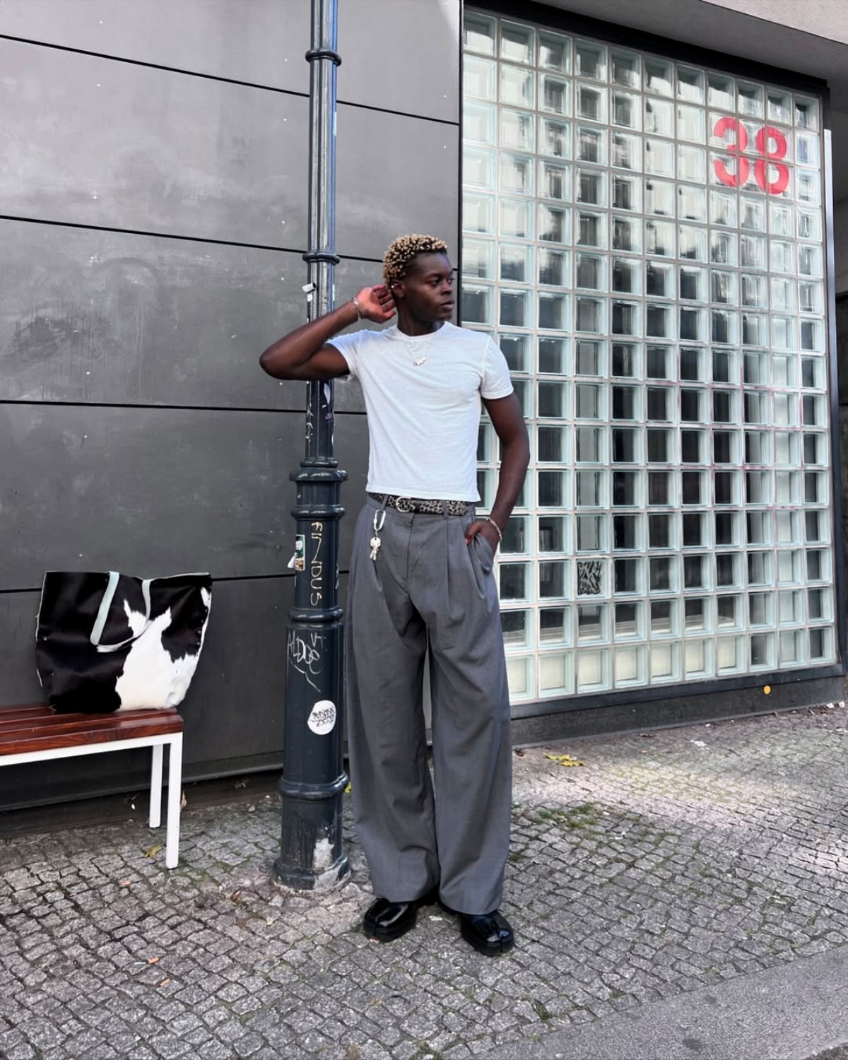 Homem negro posa para foto. Com seu braço direito apoiando sua cabeça, o rapaz olha para o horizonte vestindo uma camisa branca e calça social cinza