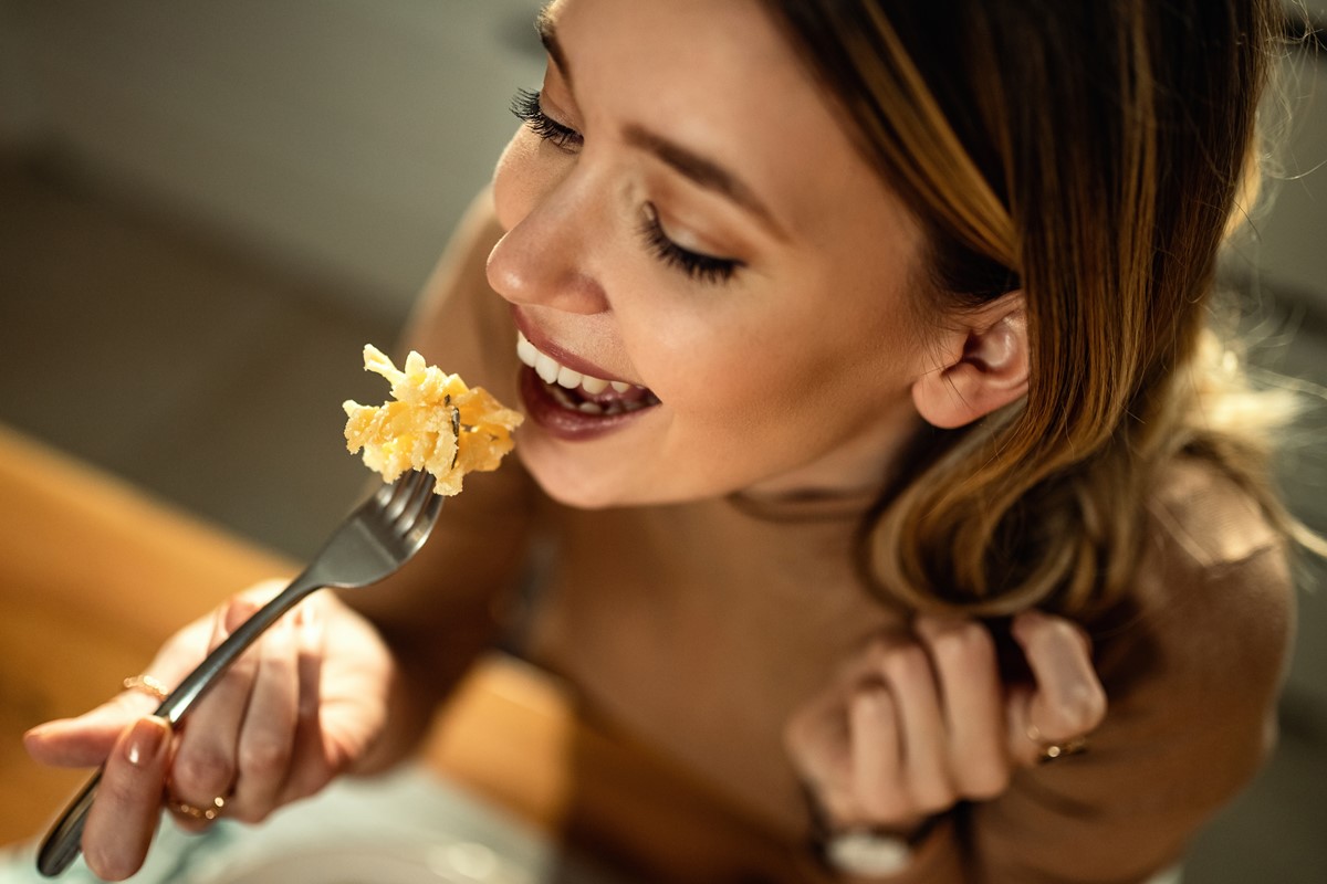 Foto colorida de mulher comendo macarrão em um garfo - Metrópoles