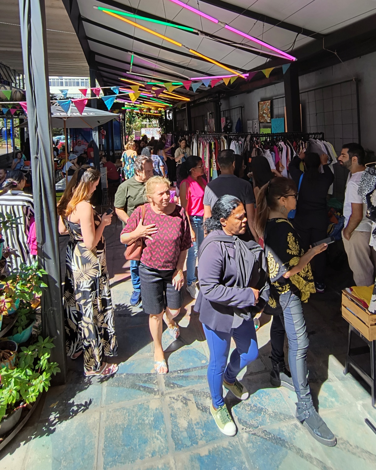 A imagem mostra uma feira de roupas com diversas pessoas caminhando pelo espaço