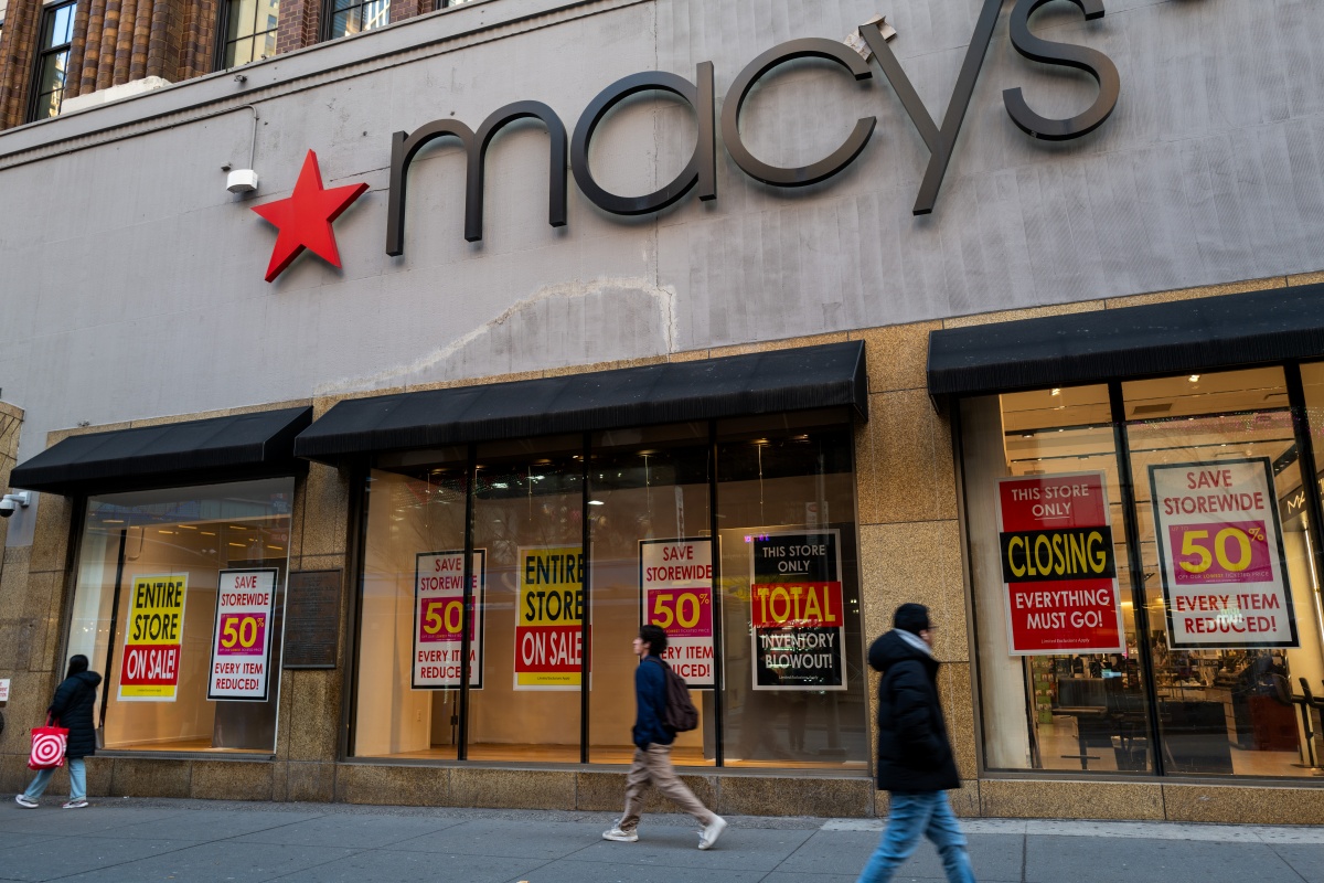 A imagem mostra as portas da loja Macy's com diversos cartazes anunciando promoções na frente do estabelecimento