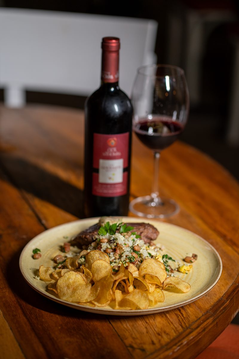 Na foto colorida, um bife ancho servido em um prato ao lado de uma taça de vinho tinto - Metrópoles 