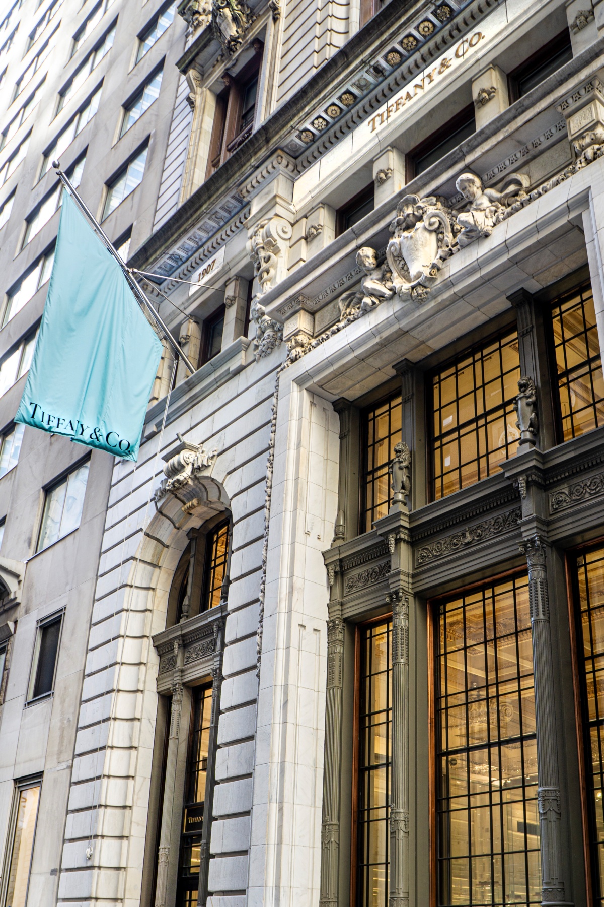 A imagem mostra a lateral de um prédio tradicional de Nova York. Presa sob a fachada, uma bandeira carrega a logo e cor da marca Tiffany & Co