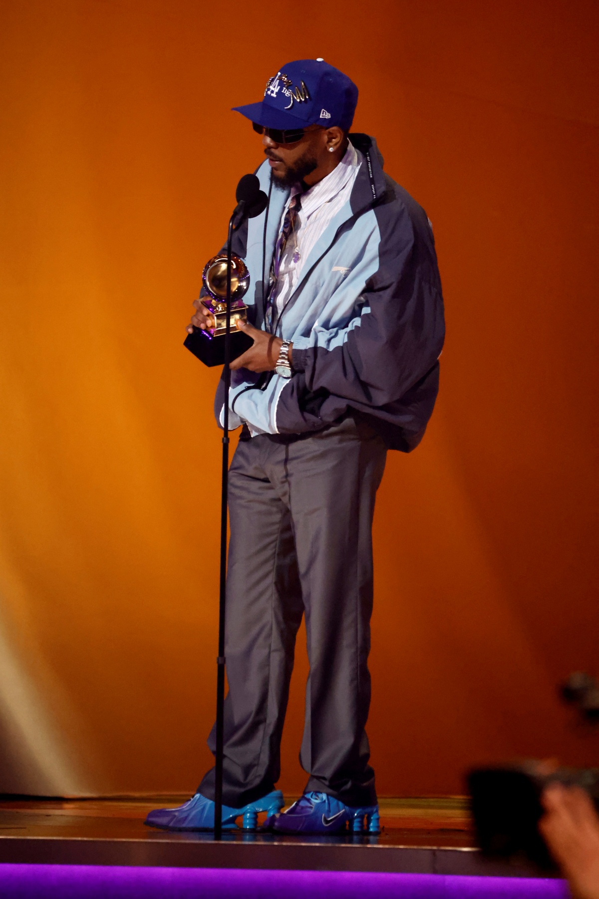 A imagem mostra o rapper Kendrick Lamar segurando um troféu enquanto fala em um microfone em frente um fundo laranja