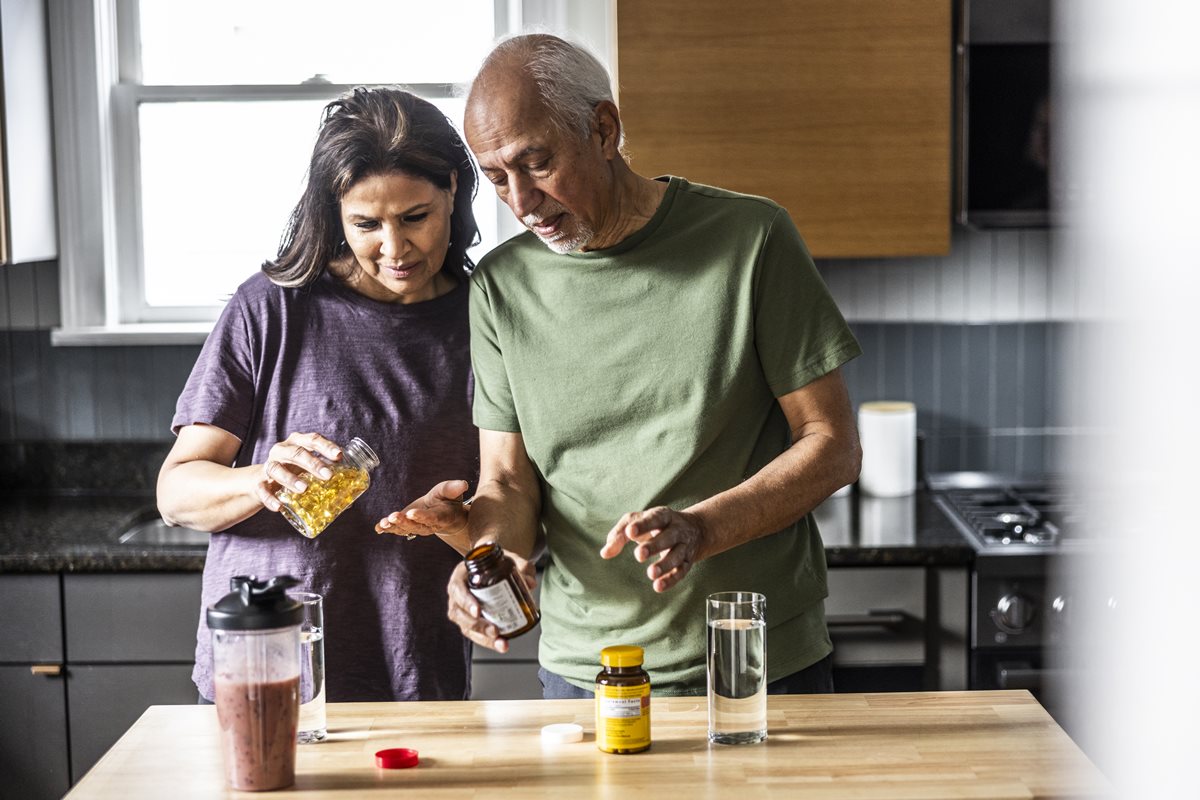 Foto colorida de mulher e homem com embalagens de suplementos. Eles estão em uma cozinha - Metrópoles