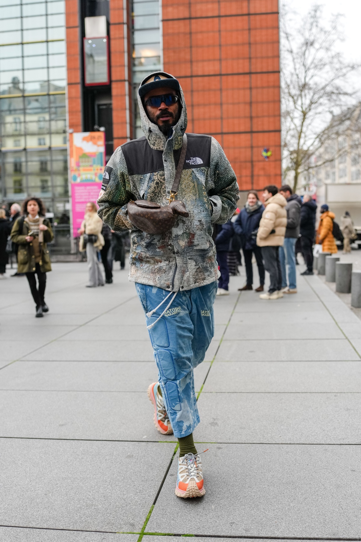 A imagem mostra um homem caminhando vestindo uma calça azul e um casaco muiticolorido com divisão preta no ombro